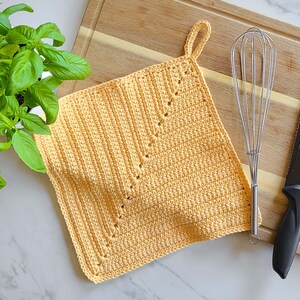 May include: A handmade, square, yellow crochet potholder with a loop for hanging. The potholder features a decorative stitch pattern and is shown on a wooden cutting board with a whisk and knife.