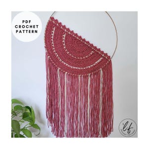 A finished sample of this crochet wall hanging pattern hung on a white wall with a plant in the corner. The crochet wall hanging is made in pink and white.