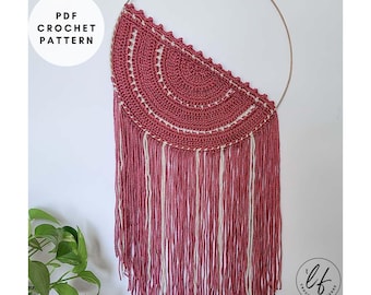 May include: A handmade crochet wall hanging in shades of burgundy and cream. The half-circle design features intricate crochet work and long fringe. The text "PDF CROCHET PATTERN" is in a white circle. A gold metal ring is at the top.