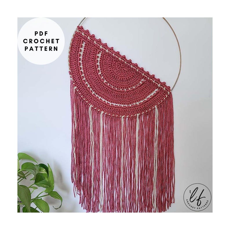 A finished sample of this crochet wall hanging pattern hung on a white wall with a plant in the corner. The crochet wall hanging is made in pink and white.
