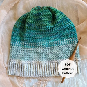 May include: A crochet hat with a green, blue and white colour gradient. The hat has a ribbed brim and is made with a textured stitch. The hat is lying on a light pink fabric with a wooden crochet hook next to it. The text "PDF Crochet Pattern" is on a white circle.