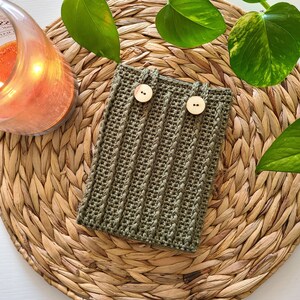 A green crochet kindle cover with two wooden buttons. The pouch is sitting on a woven wicker surface.