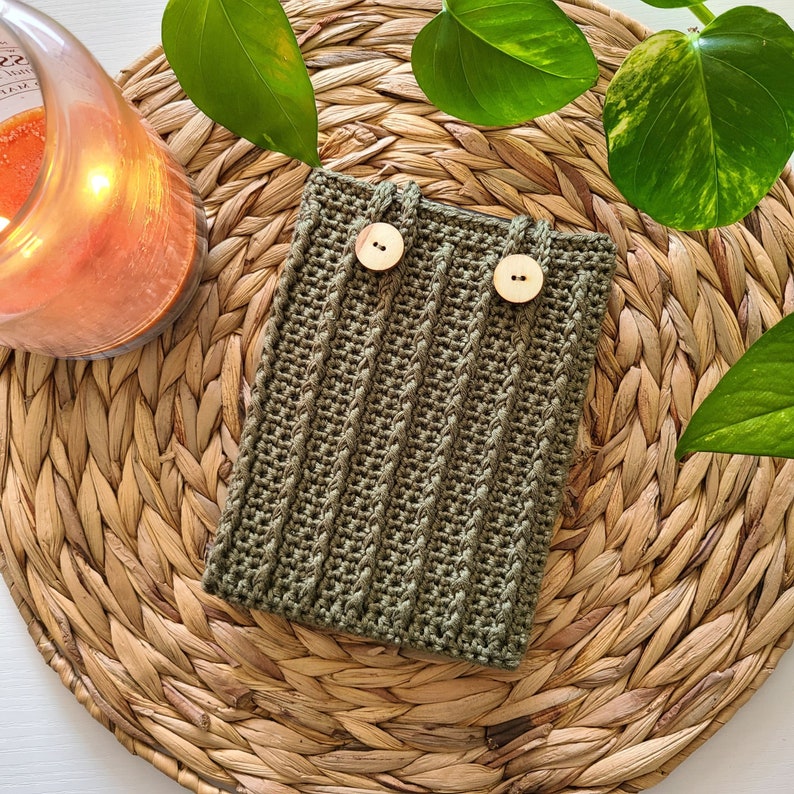 A green crochet kindle cover with two wooden buttons. The pouch is sitting on a woven wicker surface.