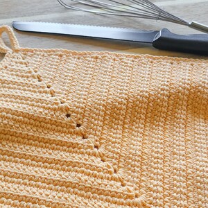 May include: Close-up of a peach-colored, handmade crochet potholder with a loop for hanging. A stainless steel knife and whisk are in the background, suggesting a kitchen setting. The potholder is made of a textured yarn.