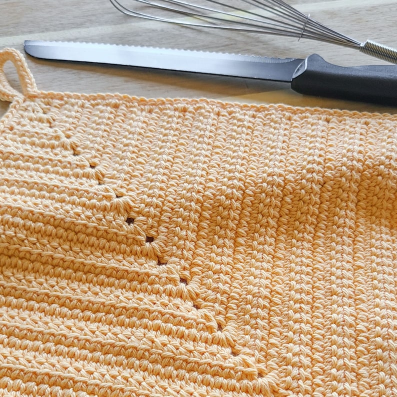 May include: Close-up of a peach-colored, handmade crochet potholder with a loop for hanging. A stainless steel knife and whisk are in the background, suggesting a kitchen setting. The potholder is made of a textured yarn.