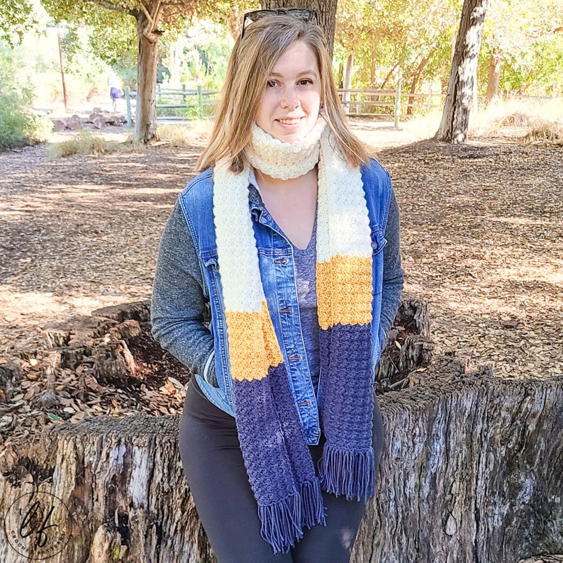 This image shows a woman wearing the sample made from this easy crochet scarf pattern. She is sitting on a large stump with a wooden background. The color block scarf is made in blue, yellow and white.