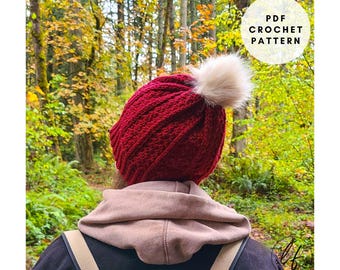 May include: A burgundy crochet hat with a cream-colored pom-pom. The hat is ribbed and has a soft texture. The image also includes the text "PDF Crochet Pattern" in a white circle.