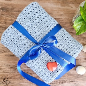 This image shows the sample of this crossed double crochet baby blanket pattern folded and wrapped in a ribbon like a present, laying flat on a wooden table.