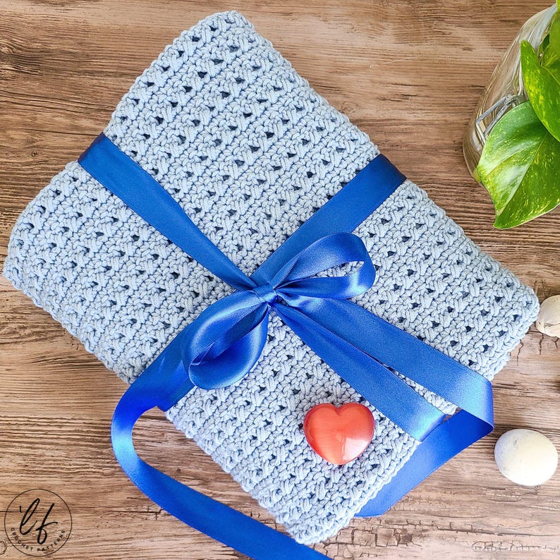 This image shows the sample of this crossed double crochet baby blanket pattern folded and wrapped in a ribbon like a present, laying flat on a wooden table.