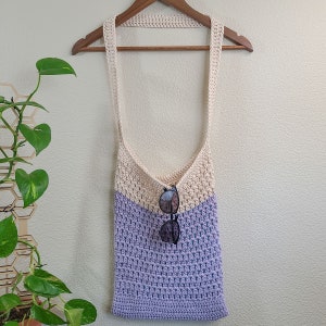 This image shows a finished bag from this crochet tote bag pattern. It is made in lavender and cream and is hanging from a clothes hanger with sunglasses hanging from the top of the bag.