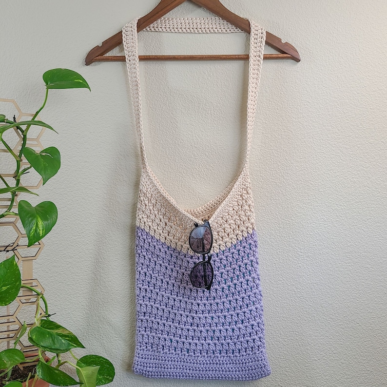 This image shows a finished bag from this crochet tote bag pattern. It is made in lavender and cream and is hanging from a clothes hanger with sunglasses hanging from the top of the bag.