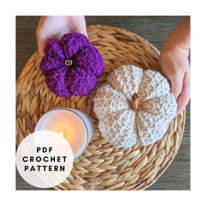 May include: Two handmade crochet pumpkins, one purple and one cream, rest on a woven tray. A cinnamon stick serves as the stem for the cream pumpkin. A lit candle sits in a silver container. The text "PDF CROCHET PATTERN" is in a white circle.
