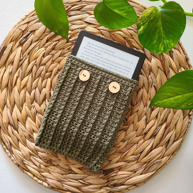 A green crocheted e-reader cover with two wooden buttons. The cover is sitting on a woven straw mat. The e-reader is visible inside the cover and displays text from a book.