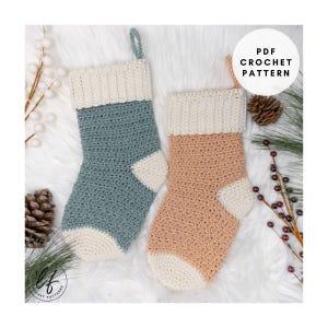 May include: Two handmade crochet Christmas stockings in blue and peach with cream-colored cuffs and toes. The image includes the text "PDF Crochet Pattern". The stockings are displayed on a white, fluffy surface with pinecones and greenery.