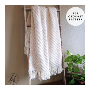Puede incluir: Manta de crochet crema con rayas diagonales y flecos, colgada de una escalera de madera. La manta tiene un aspecto texturizado. La imagen incluye el texto "PDF CROCHET PATTERN" en un círculo blanco.