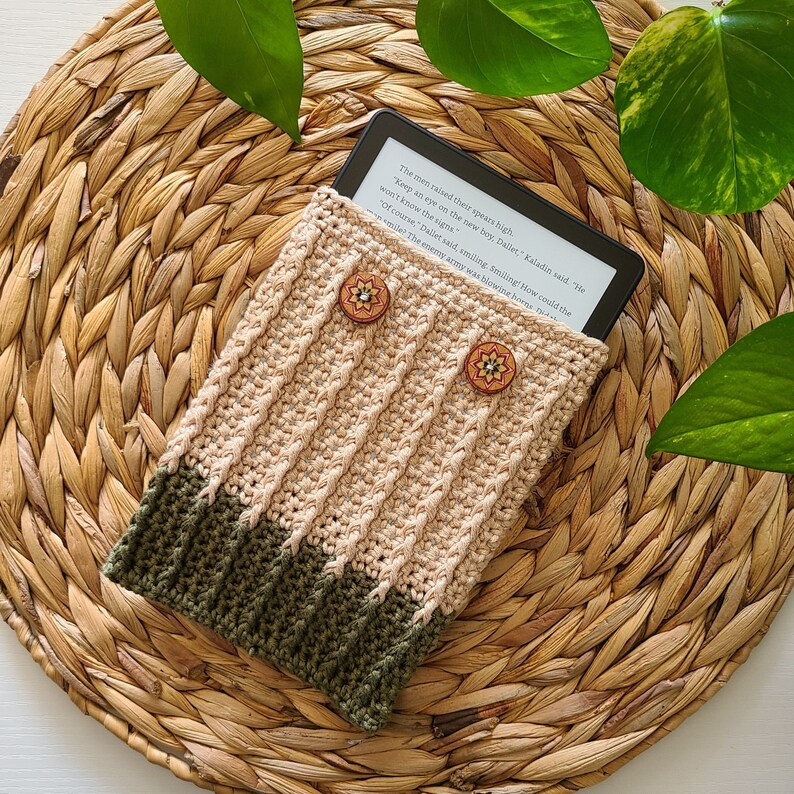 A crocheted e-reader cover in beige and green with two wooden buttons. The cover is sitting on a woven straw mat. The e-reader is visible inside the cover and displays text from a book.