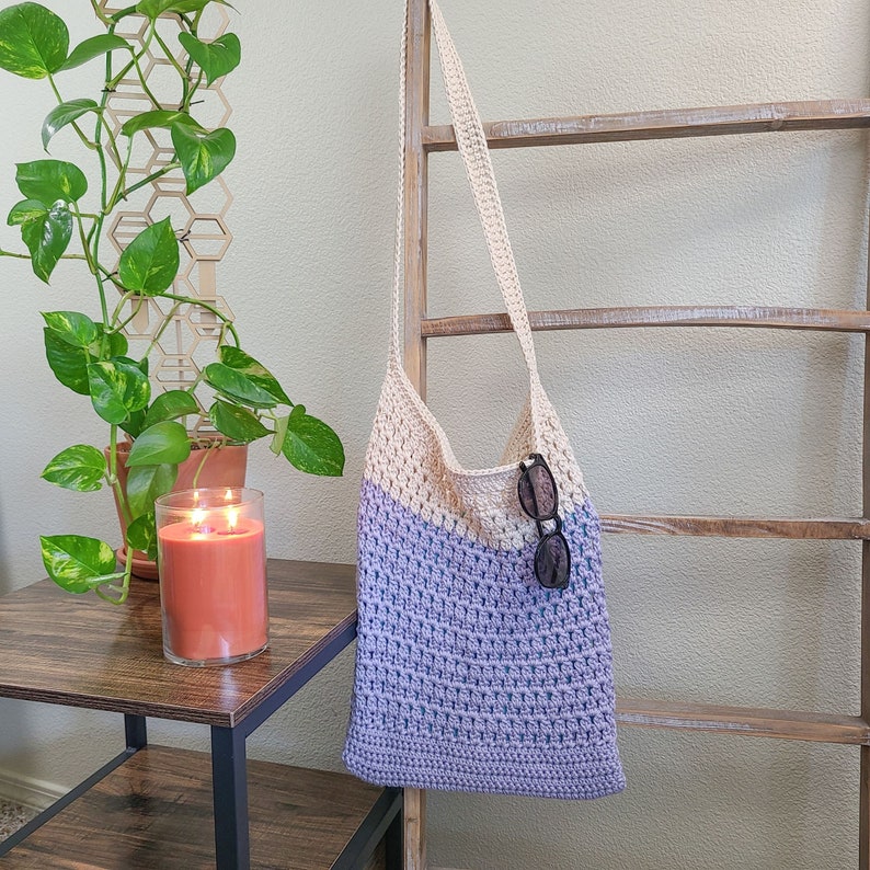 This image shows a finished bag from this crochet tote bag pattern. It is made in lavender and cream and is hanging from a blanket ladder with sunglasses hanging from the top of the bag. There is also a side table, a plant and a candle.