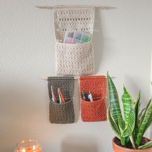 Three handmade crochet wall organizers in cream, olive green, and burnt orange hang on a wall. The cream organizer holds a clear plastic container with small craft supplies. The other two hold pens and pencils.