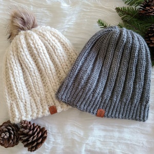 Peut inclure: Deux bonnets en tricot, un cr&egrave;me et un gris. Le bonnet cr&egrave;me a un pompon en fausse fourrure marron. Les deux ont un design en tricot c&ocirc;tel&eacute; et une petite &eacute;tiquette en cuir marron avec l'inscription "hand made with love". Des pommes de pin et des brins de conif&egrave;res sont en arri&egrave;re-plan.