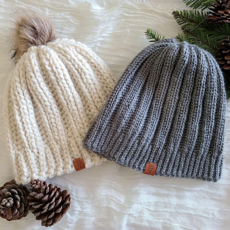 Peut inclure: Deux bonnets en tricot, un cr&egrave;me et un gris. Le bonnet cr&egrave;me a un pompon en fausse fourrure marron. Les deux ont un design en tricot c&ocirc;tel&eacute; et une petite &eacute;tiquette en cuir marron avec l'inscription "hand made with love". Des pommes de pin et des brins de conif&egrave;res sont en arri&egrave;re-plan.