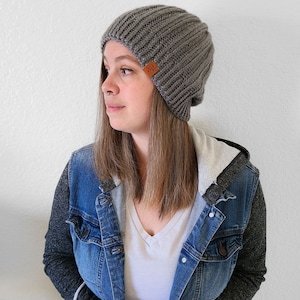 Peut inclure: Un bonnet en tricot c&ocirc;tel&eacute; gris avec une petite &eacute;tiquette en cuir marron. Le bonnet est port&eacute; avec une veste en jean et un t-shirt blanc &agrave; col en V. Un accessoire de mode pour le temps frais.