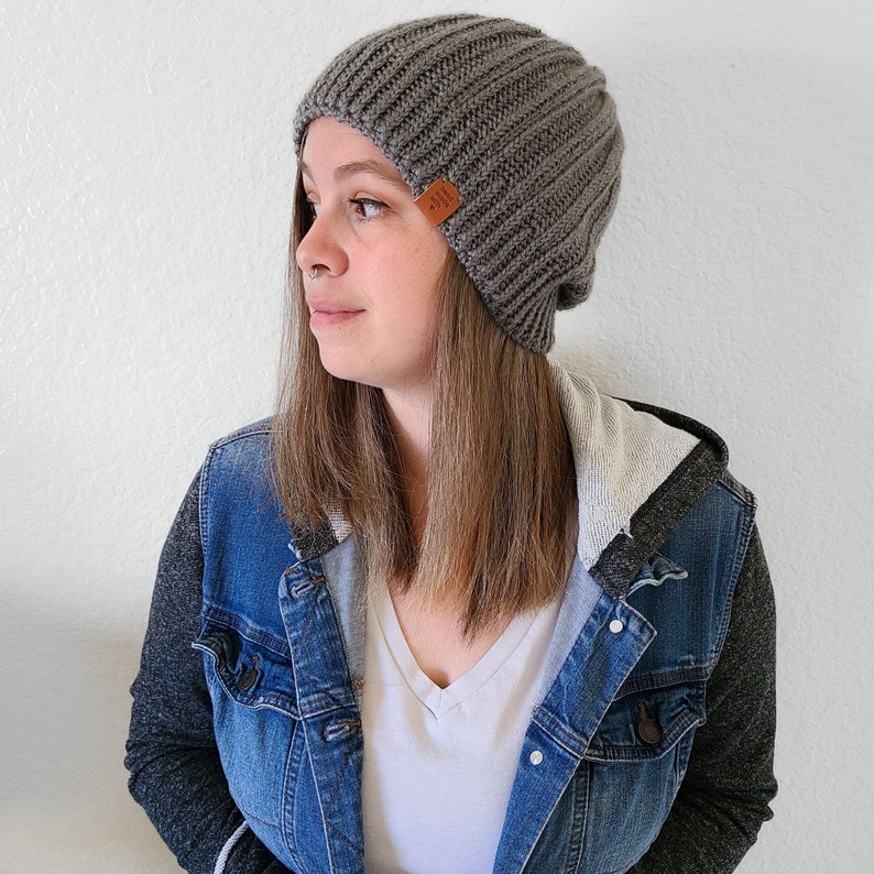 Peut inclure: Un bonnet en tricot c&ocirc;tel&eacute; gris avec une petite &eacute;tiquette en cuir marron. Le bonnet est port&eacute; avec une veste en jean et un t-shirt blanc &agrave; col en V. Un accessoire de mode pour le temps frais.