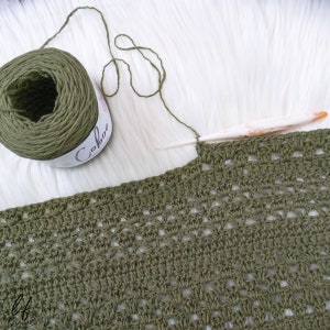 This image is a work in progress photo of the crochet triangle shawl sample. The picture is taken close up to show the detailed stitches, such as the V Stitch and Granny Stitch. Also in the picture is a half used cake of Lion Brand Coboo and a hook.