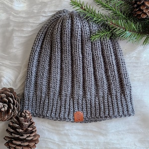 Peut inclure: Un bonnet en tricot c&ocirc;tel&eacute; gris avec une petite &eacute;tiquette marron indiquant "fait &agrave; la main avec amour". Le chapeau est pr&eacute;sent&eacute; sur une surface blanche avec des pommes de pin et une branche de conif&egrave;re.