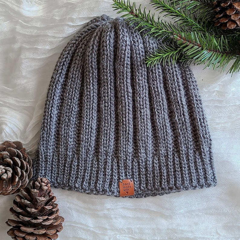 Peut inclure: Un bonnet en tricot c&ocirc;tel&eacute; gris avec une petite &eacute;tiquette marron indiquant "fait &agrave; la main avec amour". Le chapeau est pr&eacute;sent&eacute; sur une surface blanche avec des pommes de pin et une branche de conif&egrave;re.