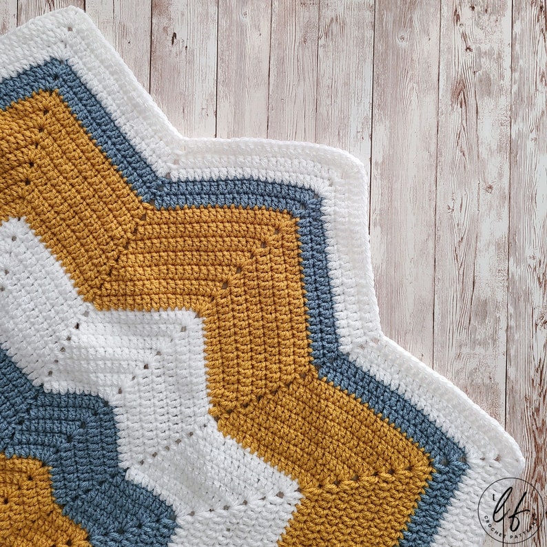 A crocheted blanket with a ripple pattern in white, mustard yellow, and blue. The blanket is made with a soft yarn and has a textured look.
