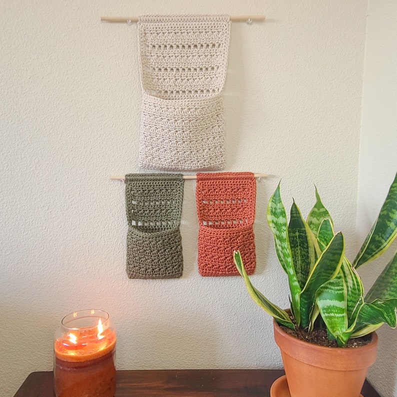 Three crochet hanging baskets in beige, olive green, and terracotta orange hang on wooden dowels. The organizers have a pocket design and are mounted on a light-colored wall. A potted snake plant and a lit candle are in the foreground.