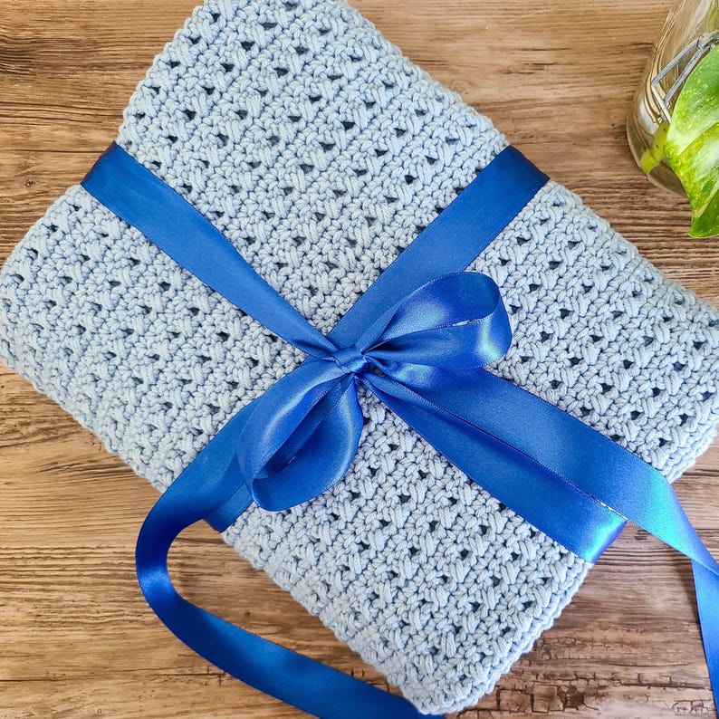This image shows the sample of this crossed double crochet baby blanket pattern folded and wrapped in a ribbon like a present, laying flat on a wooden table.