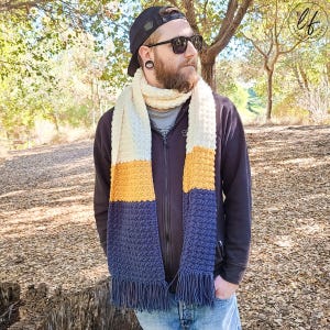 This image shows a man wearing the sample made from this easy crochet mens scarf pattern with a wooden background. The crochet scarf is made in blue, yellow and white.