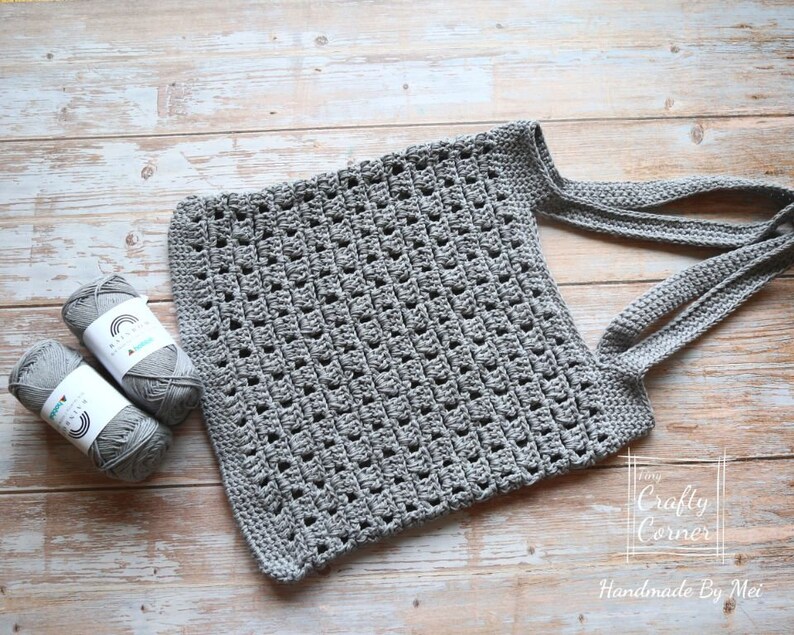 K&ouml;nnte beinhalten: Eine graue geh&auml;kelte Einkaufstasche mit zwei Henkeln. Die Tasche ist mit einem einfachen Stichmuster gearbeitet und hat eine quadratische Form. Zwei Kn&auml;uel graues Garn befinden sich im Vordergrund.
