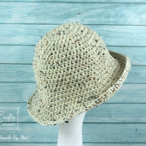 May include: A crocheted bucket hat in a speckled cream color with a black trim. The hat is displayed on a white mannequin against a light blue wooden background.