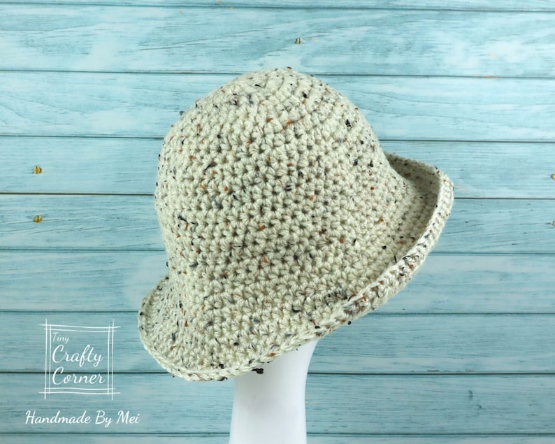 May include: A crocheted bucket hat in a speckled cream color with a black trim. The hat is displayed on a white mannequin against a light blue wooden background.