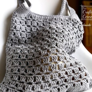 K&ouml;nnte beinhalten: Eine graue geh&auml;kelte Einkaufstasche mit quadratischem Muster. Die Tasche liegt auf einem wei&szlig;en Stuhl. Die Tasche wurde von Tiny Crafty Corner, Handmade By Mei, hergestellt.