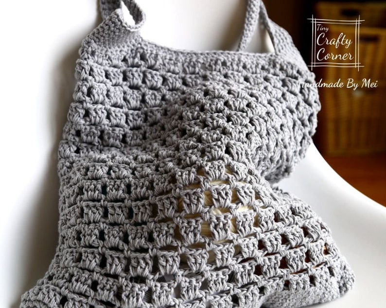 K&ouml;nnte beinhalten: Eine graue geh&auml;kelte Einkaufstasche mit quadratischem Muster. Die Tasche liegt auf einem wei&szlig;en Stuhl. Die Tasche wurde von Tiny Crafty Corner, Handmade By Mei, hergestellt.