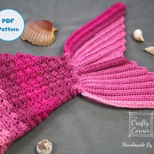 May include: A pink and red crocheted mermaid tail blanket with a scalloped edge. The blanket is made with a textured stitch and has a gradient of colour from pink to red. The blanket is lying on a grey surface with seashells.