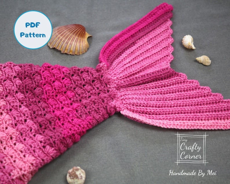 May include: A pink and red crocheted mermaid tail blanket with a scalloped edge. The blanket is made with a textured stitch and has a gradient of color from pink to red. The blanket is lying on a gray surface with seashells.