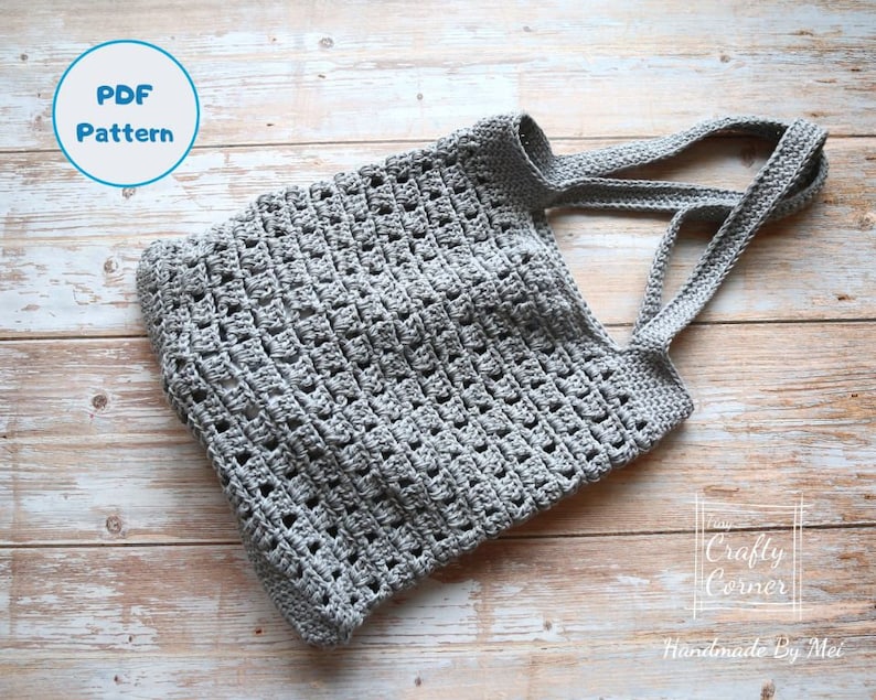 K&ouml;nnte beinhalten: Eine graue geh&auml;kelte Einkaufstasche mit breiten Henkeln. Die Tasche ist mit einem einfachen Stichmuster gearbeitet und hat einen l&auml;ssigen, ungezwungenen Look. Das Bild enth&auml;lt den Text "PDF Pattern" und "The Crafty Corner Handmade By Mei".