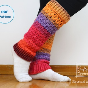 Puede incluir: Un par de polainas de crochet de colores con un patrón a rayas en tonos de morado, naranja, rojo y marrón. Las polainas se llevan sobre calcetines blancos y se muestran sobre un fondo de madera. El texto "Crafty Corner Handmade By Mei" es visible en la esquina inferior derecha de la imagen.
