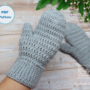 May include: A pair of gray crocheted mittens with a textured stitch pattern. The mittens are shown on a wooden surface with a green plant in the background. The text "PDF Pattern" is in a circle at the top of the image. The text "Crafty Corner Handmade By Mei" is at the bottom of the image.