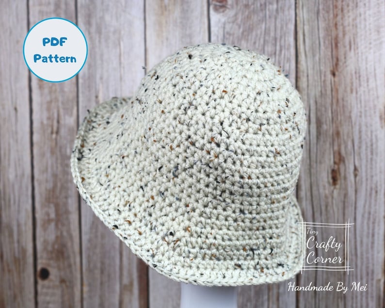 May include: A crocheted bucket hat in a speckled beige color. The hat is on a wooden background with a blue circle that says "PDF Pattern" in white text.