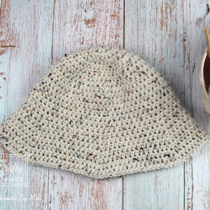 May include: A crocheted bucket hat in a speckled cream color with brown and black flecks. The hat is lying flat on a light blue and white wood surface.