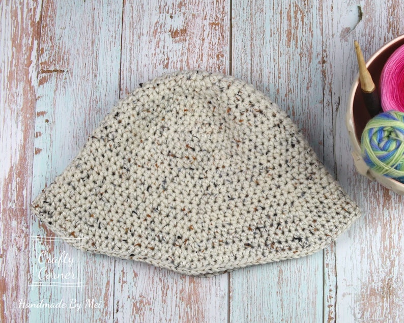 May include: A crocheted bucket hat in a speckled cream color with brown and black flecks. The hat is lying flat on a light blue and white wood surface.