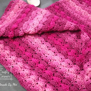 May include: A pink and purple crocheted blanket with a textured pattern. The blanket is folded in half and the text "Tiny Crafty Corner Handmade By Mei" is visible in the bottom left corner.