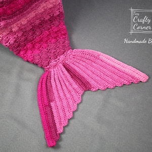 May include: A pink crocheted mermaid tail blanket with a scalloped edge. The blanket is made with a gradient of pink yarn, starting with a darker pink at the top and fading to a lighter pink at the bottom. The tail is crocheted in a textured stitch, giving it a dimensional look. The blanket is handmade by Tiny Crafty Corner.