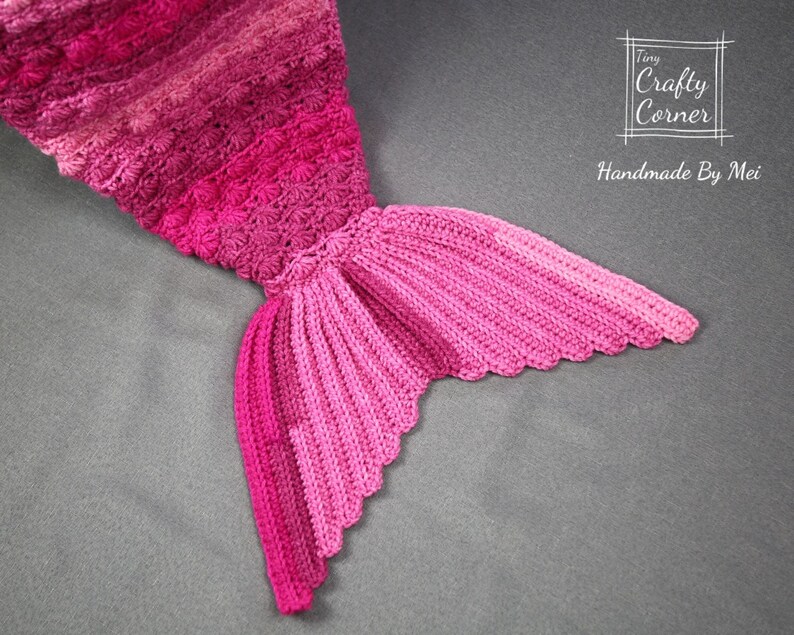 May include: A pink crocheted mermaid tail blanket with a scalloped edge. The blanket is made with a gradient of pink yarn, starting with a darker pink at the top and fading to a lighter pink at the bottom. The tail is crocheted in a textured stitch, giving it a dimensional look. The blanket is handmade by Tiny Crafty Corner.
