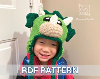 May include: A child wearing a green and white crocheted dinosaur hat. The hat has a large, round head with a long snout and two horns. The hat also has two large, black eyes and a small, white nose. The hat is being worn by a child with a big smile. The text "PDF PATTERN" is printed in blue letters on a white background.